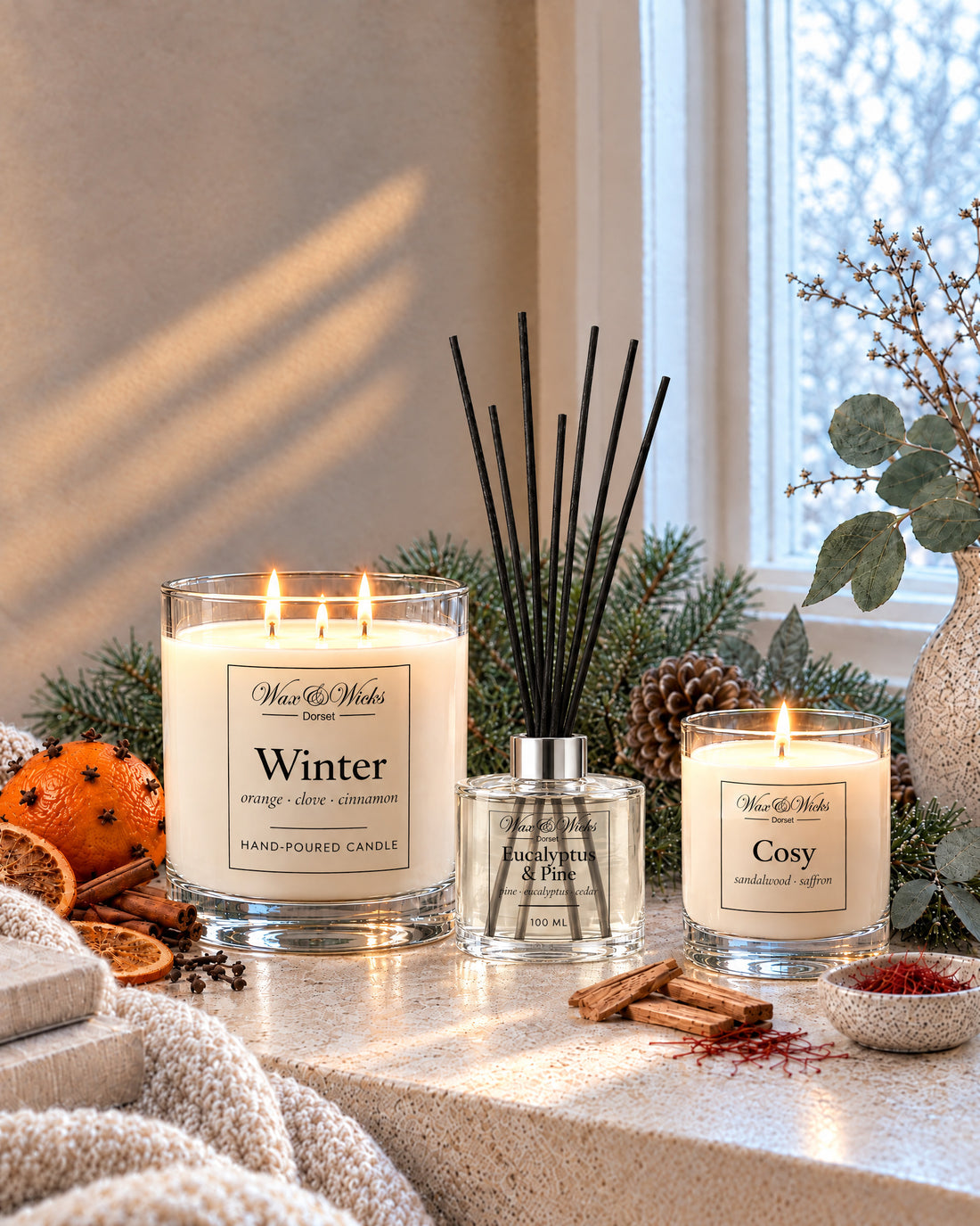 A large Winter candle, a Eucalyptus & Pine reed diffuser and a small Cosy candle, lit, by a snowy window with a clove-studded orange, dried orange slices, pine, eucalyptus and a knitted throw