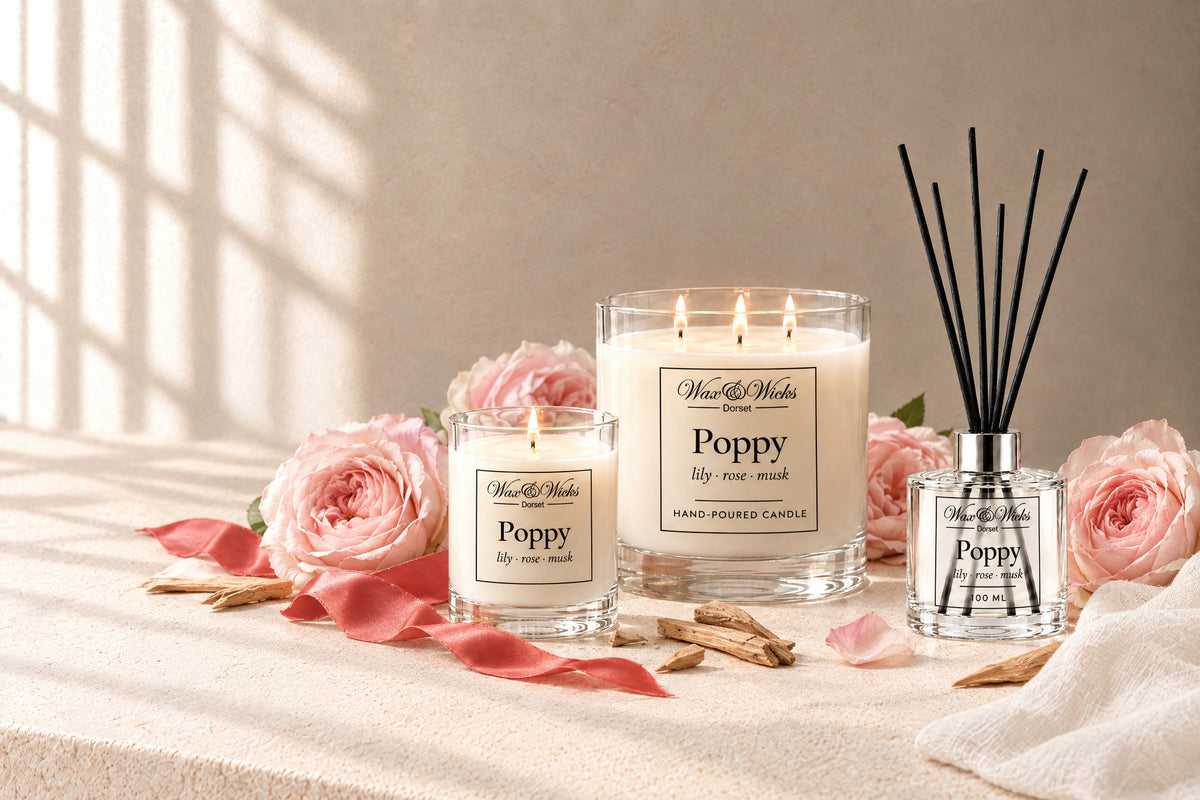 Poppy small and large candles and reed diffuser together, styled with pale pink garden roses, sandalwood chips, white muslin and a coral-red ribbon