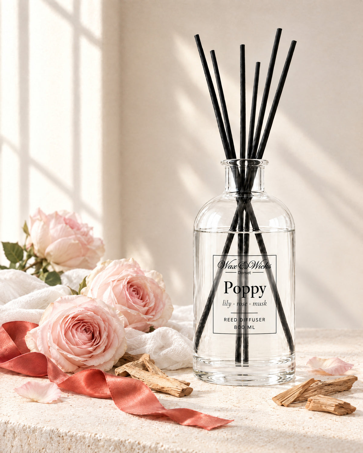 Poppy 800 ml large reed diffuser in apothecary-style glass with extra-long reeds, styled with pale pink garden roses, sandalwood chips, white muslin and a coral-red ribbon