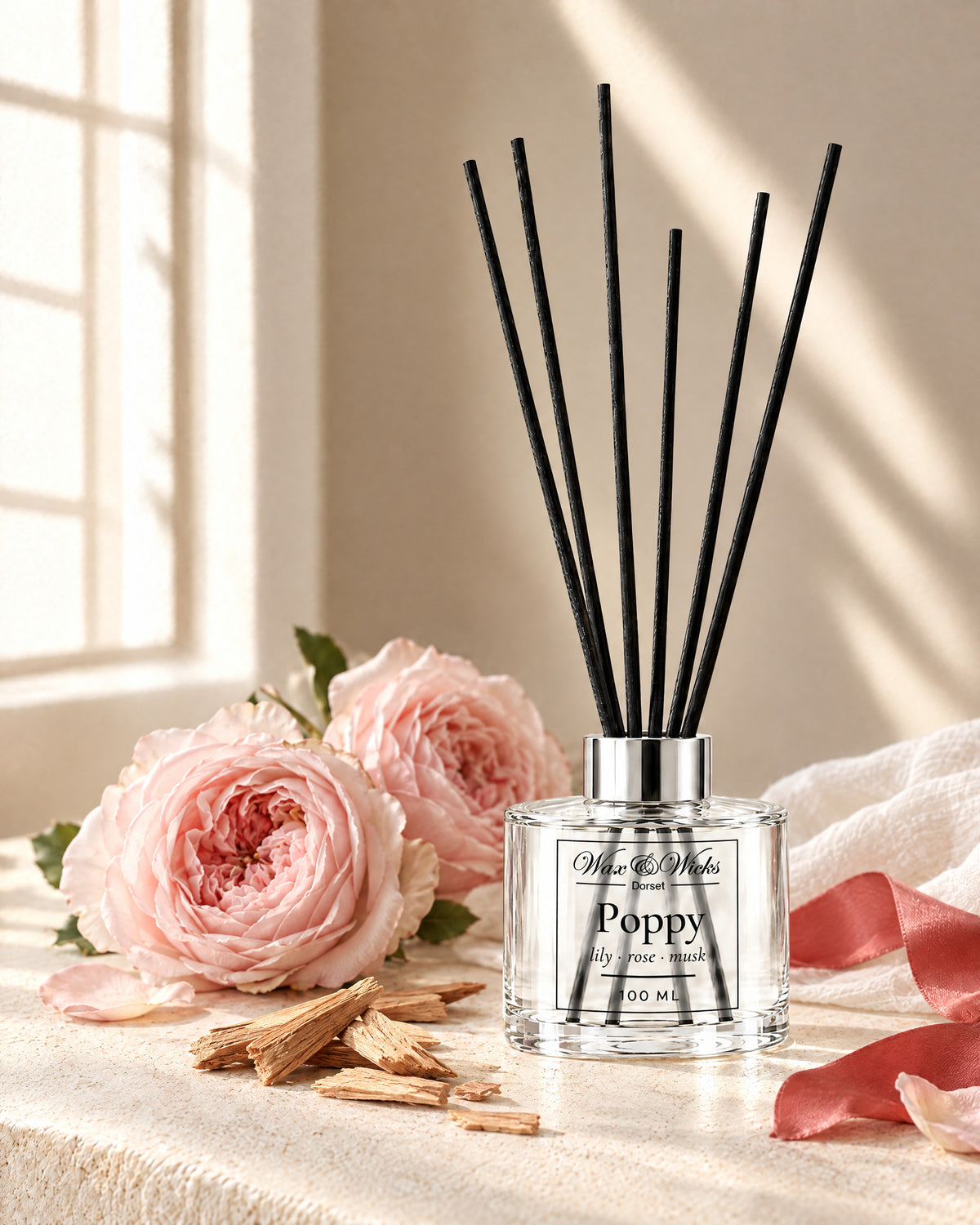 Poppy 100 ml reed diffuser with black fibre reeds, styled with pale pink garden roses, sandalwood chips, white muslin and a coral-red ribbon