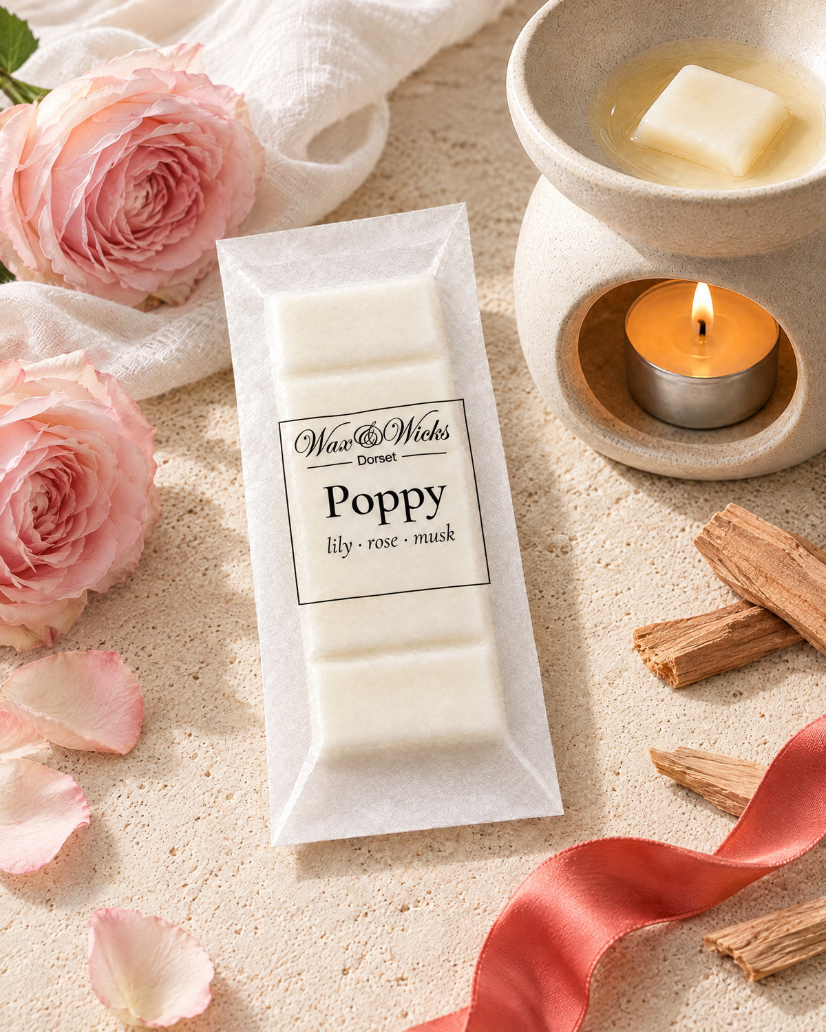 Poppy wax melt snap bar, beside a tea-light wax burner, styled with pale pink garden roses, sandalwood chips, white muslin and a coral-red ribbon