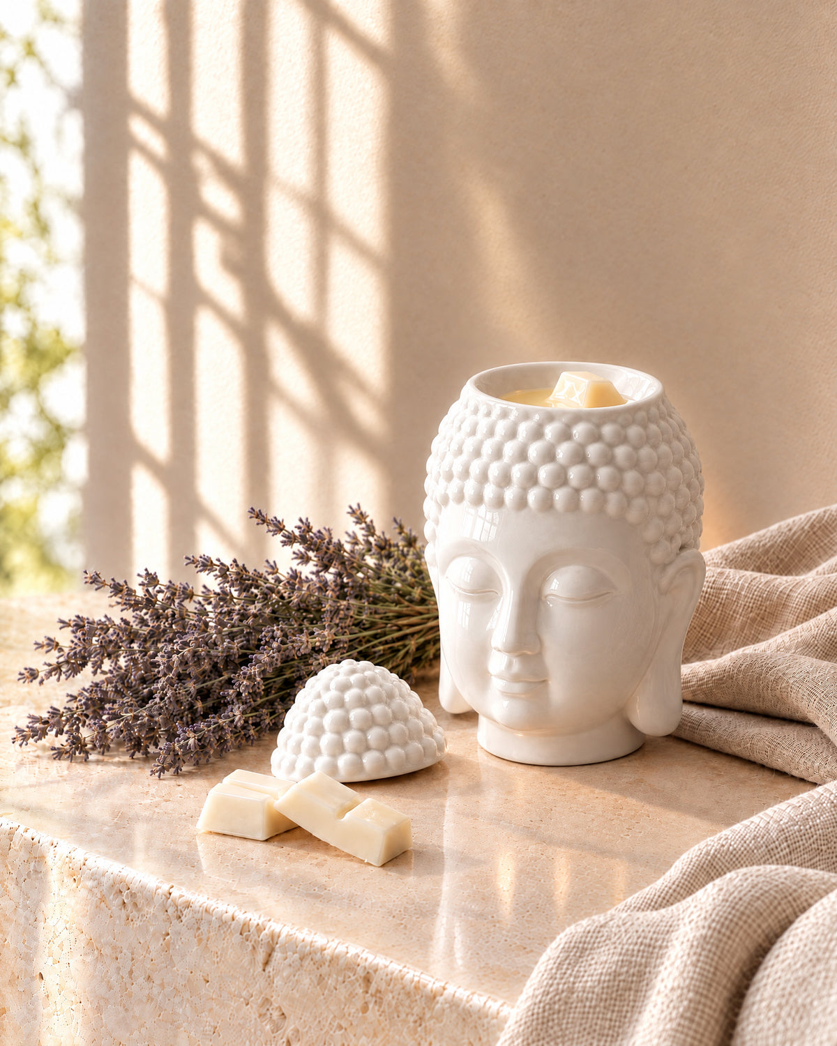White Prince Buddha wax melt burner with its top-knot lid lifted off and wax melting in the dish, beside wax melt segments, dried lavender and linen on a stone ledge