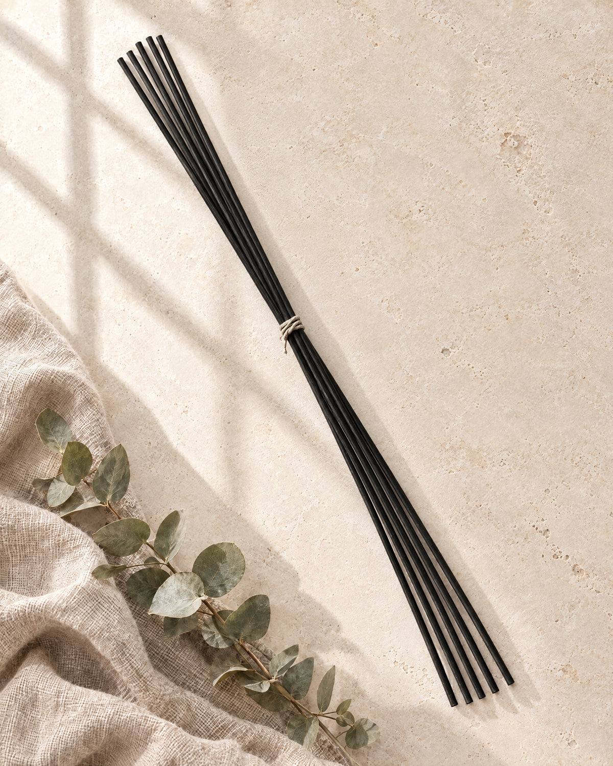Extra-long black fibre replacement reeds tied with natural cord, lying on stone with linen and a sprig of eucalyptus