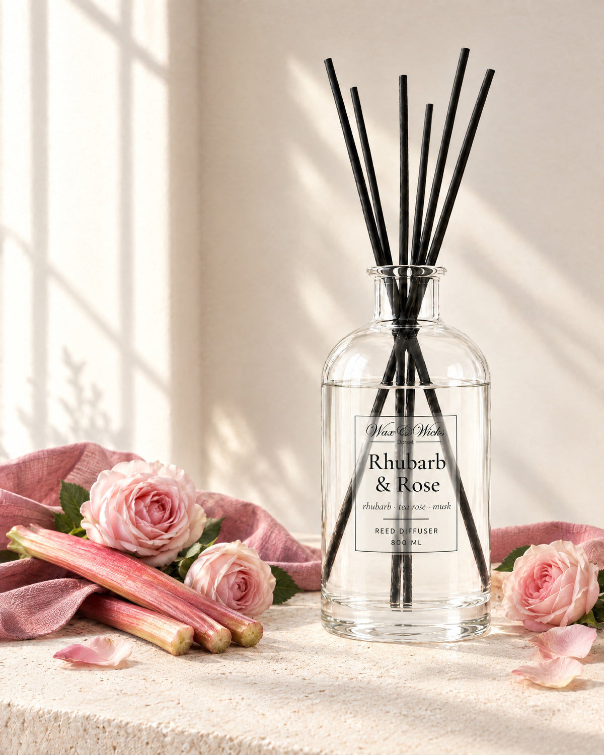 Rhubarb & Rose 800 ml large reed diffuser in apothecary-style glass with extra-long reeds, styled with stalks of pink rhubarb, pale pink roses and rose petals, with a dusky pink linen