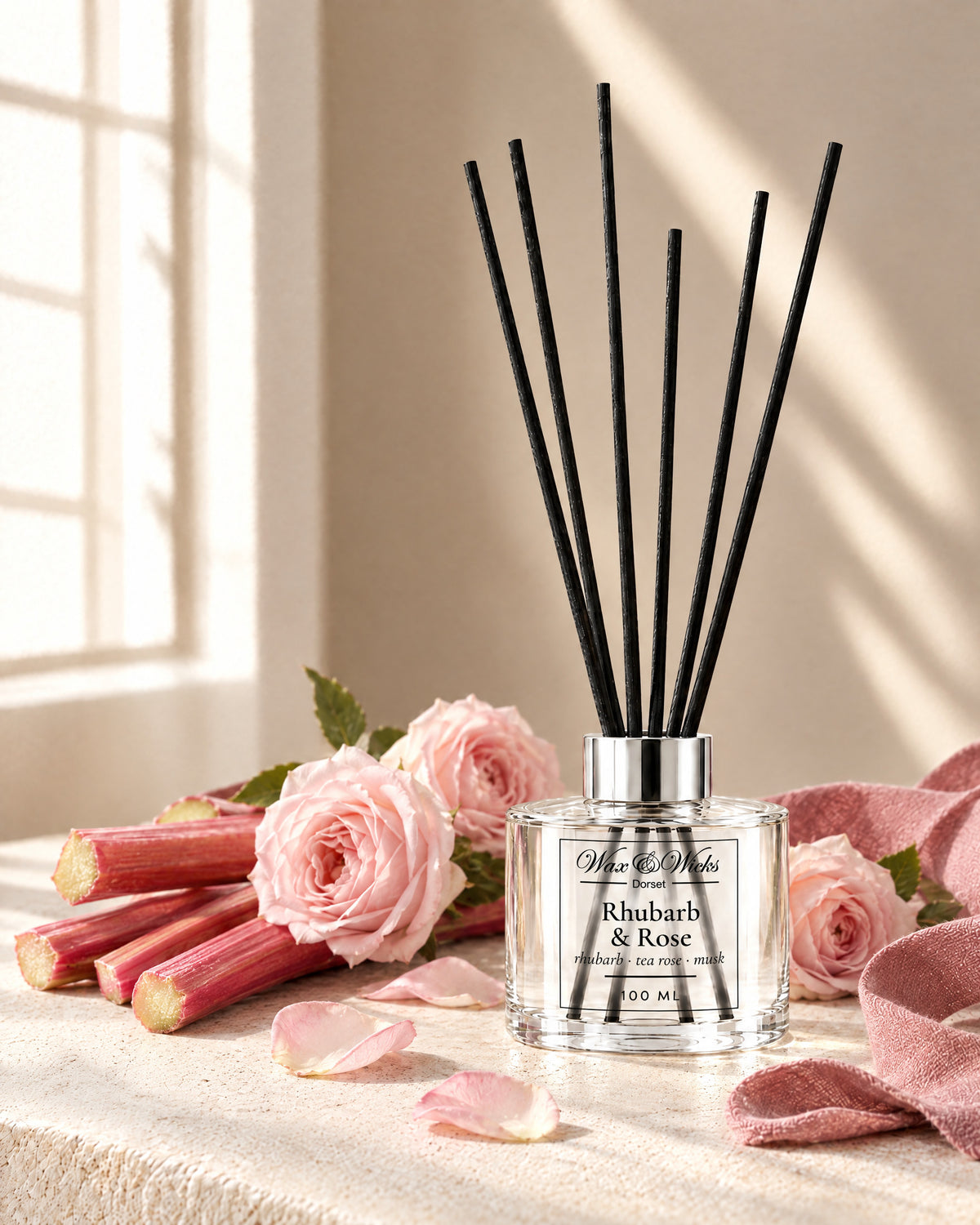 Rhubarb & Rose 100 ml reed diffuser with black fibre reeds, styled with stalks of pink rhubarb, pale pink roses and rose petals, with a dusky pink linen