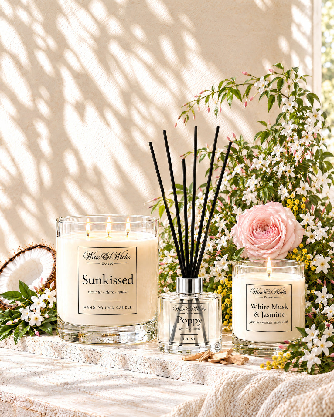 A large Sunkissed candle, a Poppy reed diffuser and a small White Musk & Jasmine candle, lit, with half a coconut, sprays of white jasmine, a pink rose, mimosa and sandalwood in warm summer light