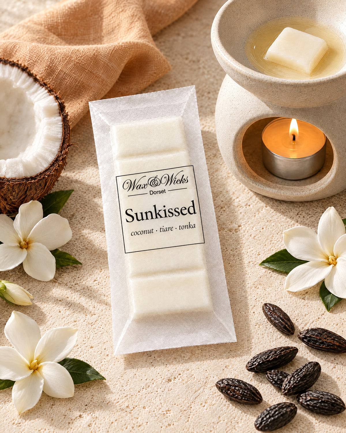Sunkissed wax melt snap bar, beside a tea-light wax burner, styled with half a coconut, white tiare flowers and tonka beans, with a peach linen