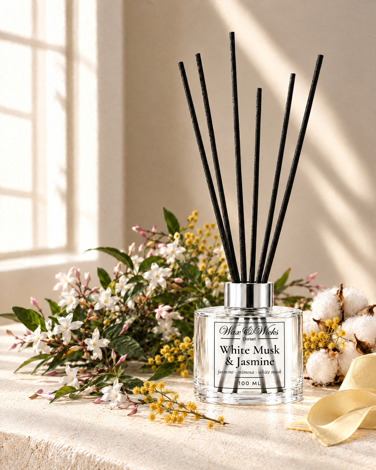 White Musk & Jasmine 100 ml reed diffuser with black fibre reeds, styled with sprays of white jasmine, yellow mimosa and cotton flowers, with a pale yellow ribbon
