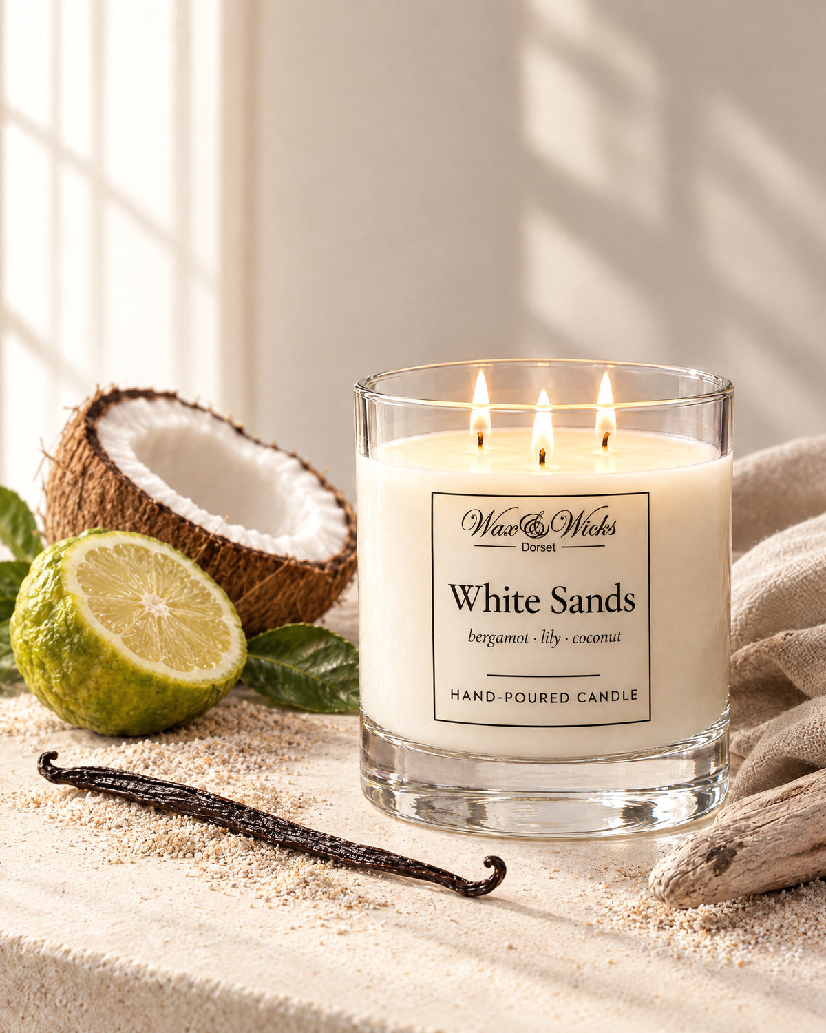 Large White Sands candle, three wicks, in a clear glass jar, styled with half a bergamot, half a coconut, a vanilla pod and driftwood on pale sand