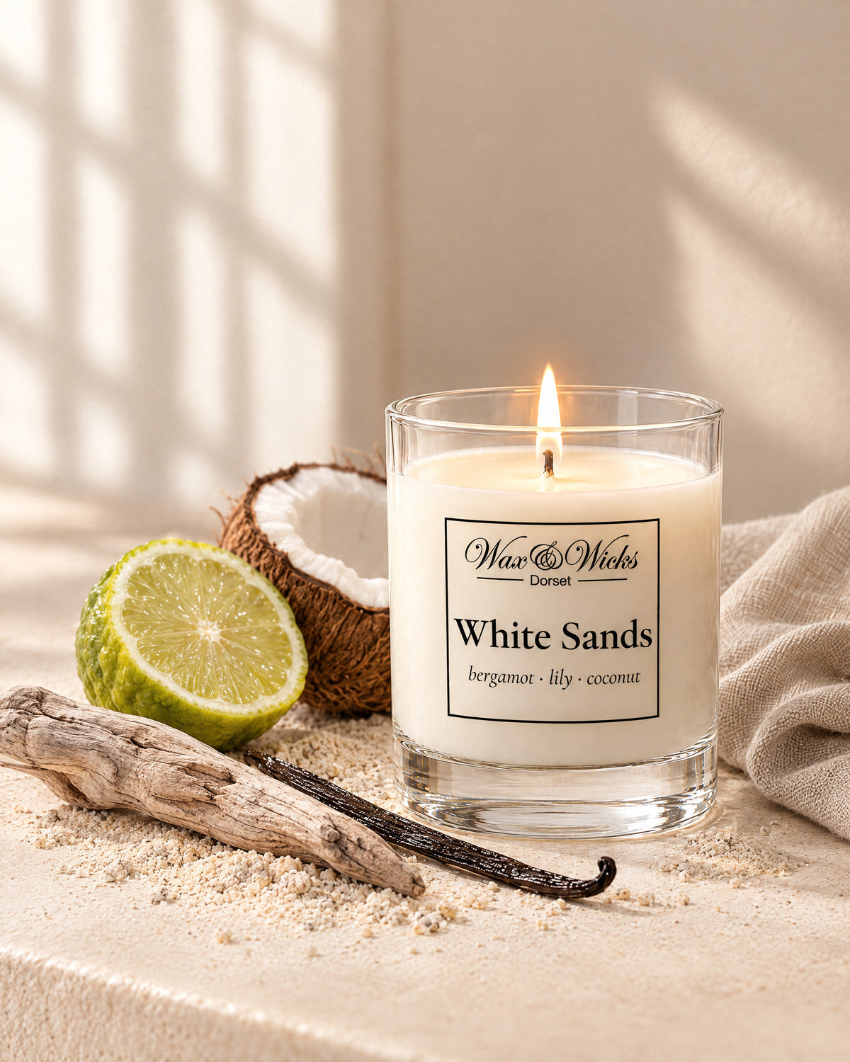 Small White Sands candle, one wick, in a clear glass jar, styled with half a bergamot, half a coconut, a vanilla pod and driftwood on pale sand