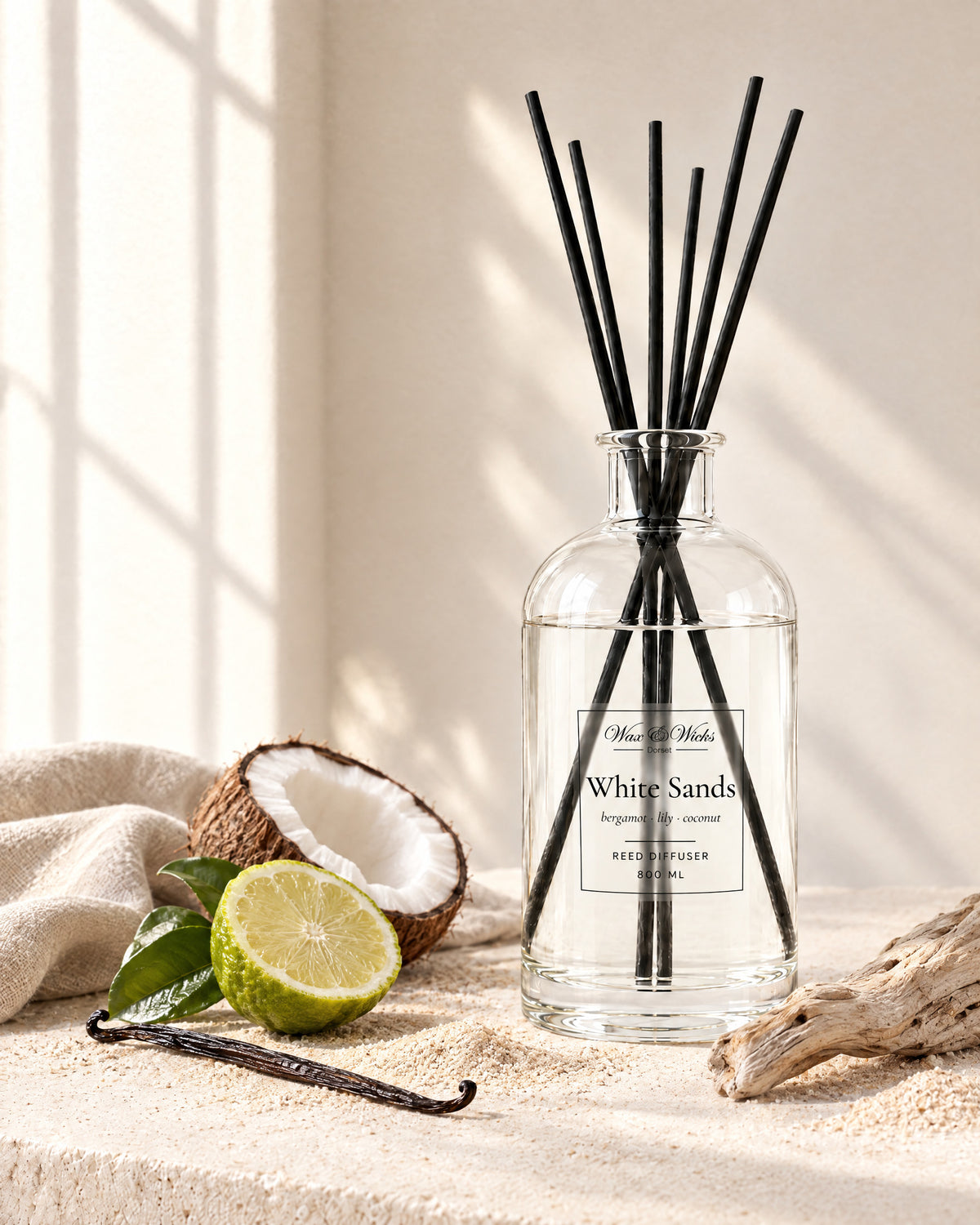 White Sands 800 ml large reed diffuser in apothecary-style glass with extra-long reeds, styled with half a bergamot, half a coconut, a vanilla pod and driftwood on pale sand