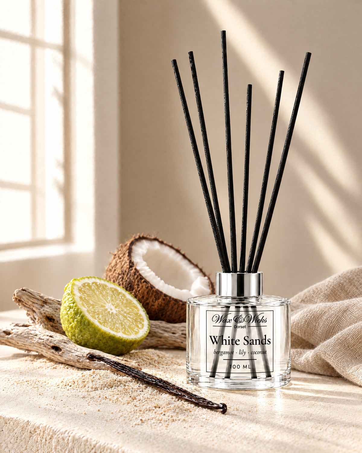 White Sands 100 ml reed diffuser with black fibre reeds, styled with half a bergamot, half a coconut, a vanilla pod and driftwood on pale sand