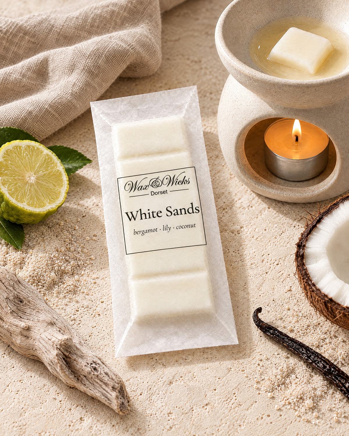White Sands wax melt snap bar, beside a tea-light wax burner, styled with half a bergamot, half a coconut, a vanilla pod and driftwood on pale sand