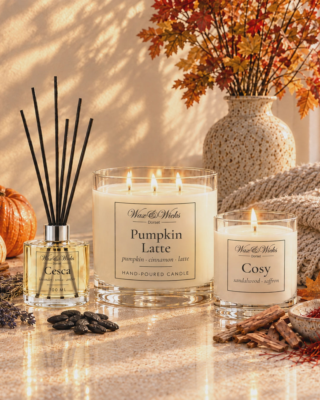 A Pumpkin Latte candle, a Cesca reed diffuser and a small Cosy candle, lit, with pumpkins, autumn leaves and a knitted throw in low golden light